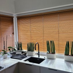 Świat Rolet i Żaluzji - Kuchnia z oknem przesłoniętym poziomymi, drewnianymi lamelami. Na parapecie doniczki z zielonymi roślinami. Widok na zlew i płytę grzewczą.