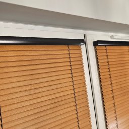 Świat Rolet i Żaluzji - Dwa okna z zamontowanymi poziomymi, drewnianymi żaluzjami w odcieniach brązu, z czarnymi elementami mocującymi. Widok z bliska, wnętrze pomieszczenia.