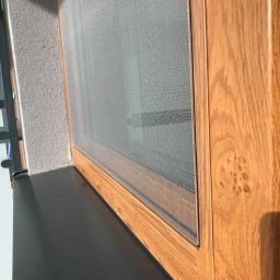 Świat Rolet i Żaluzji - Zamontowana moskitiera w oknie z drewnianą ramą, widok z boku, szara ściana w tle. Siatka moskitiery jest napięta i równa.