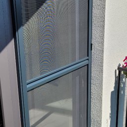 Świat Rolet i Żaluzji - Szara moskitiera ramkowa w oknie, widok z balkonu. Na ścianie widoczne cienie, a w tle marmurowa podłoga. Dyskretna ochrona przed owadami.