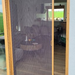 Świat Rolet i Żaluzji - Zewnętrzna moskitiera plisowana w drewnianej ramie, zamontowana na tarasie. Widok na wnętrze domu przez siatkę. Nowoczesne rozwiązanie na owady.