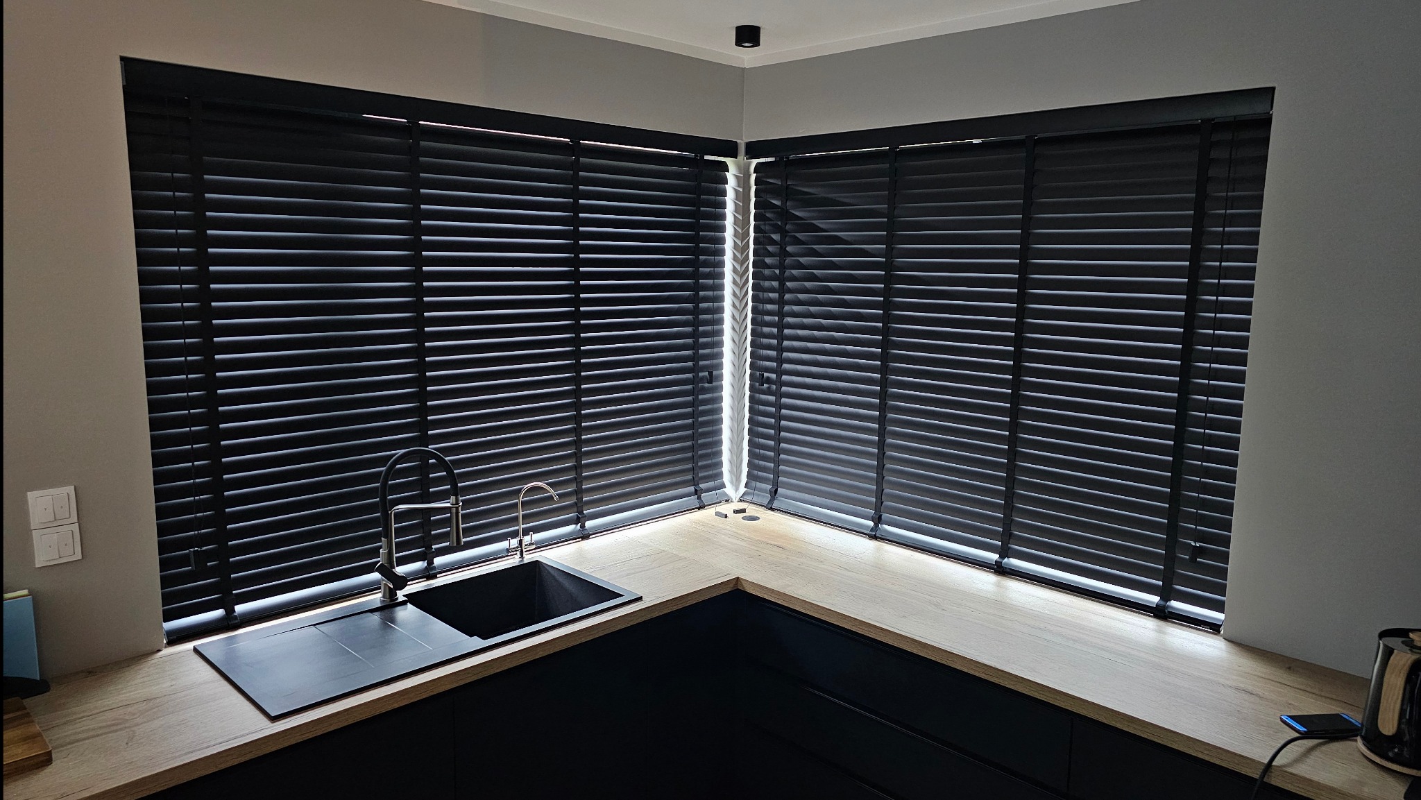 Czarne żaluzje poziome w kuchni, zamontowane w narożnym oknie nad blatem i zlewem. Nowoczesny design, szare ściany, drewniany blat. Widok z góry.
