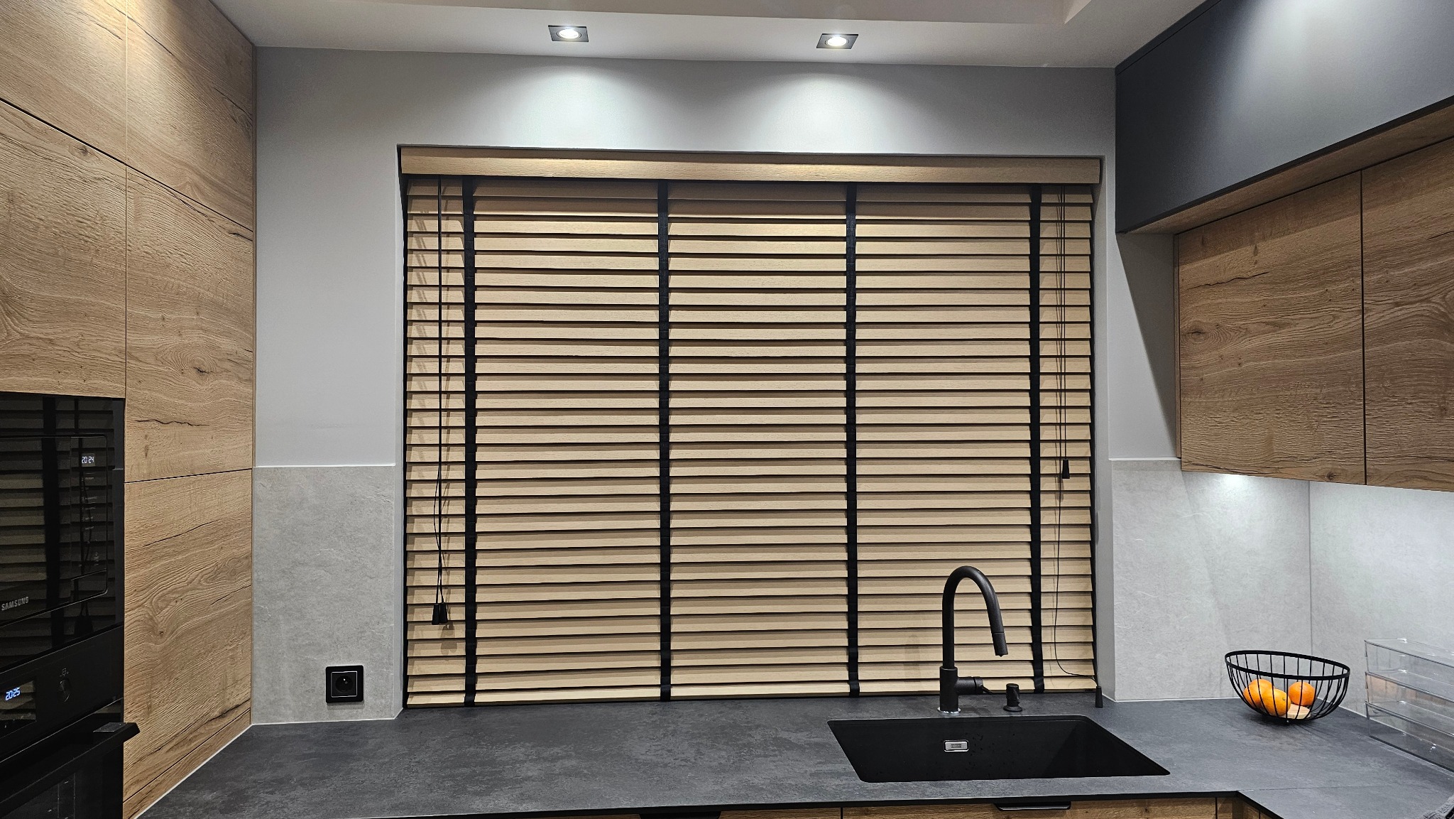 Jasne, poziome przesłony okienne z czarnymi taśmami w nowoczesnej kuchni z drewnianymi szafkami i czarnym zlewem. Oświetlenie sufitowe.