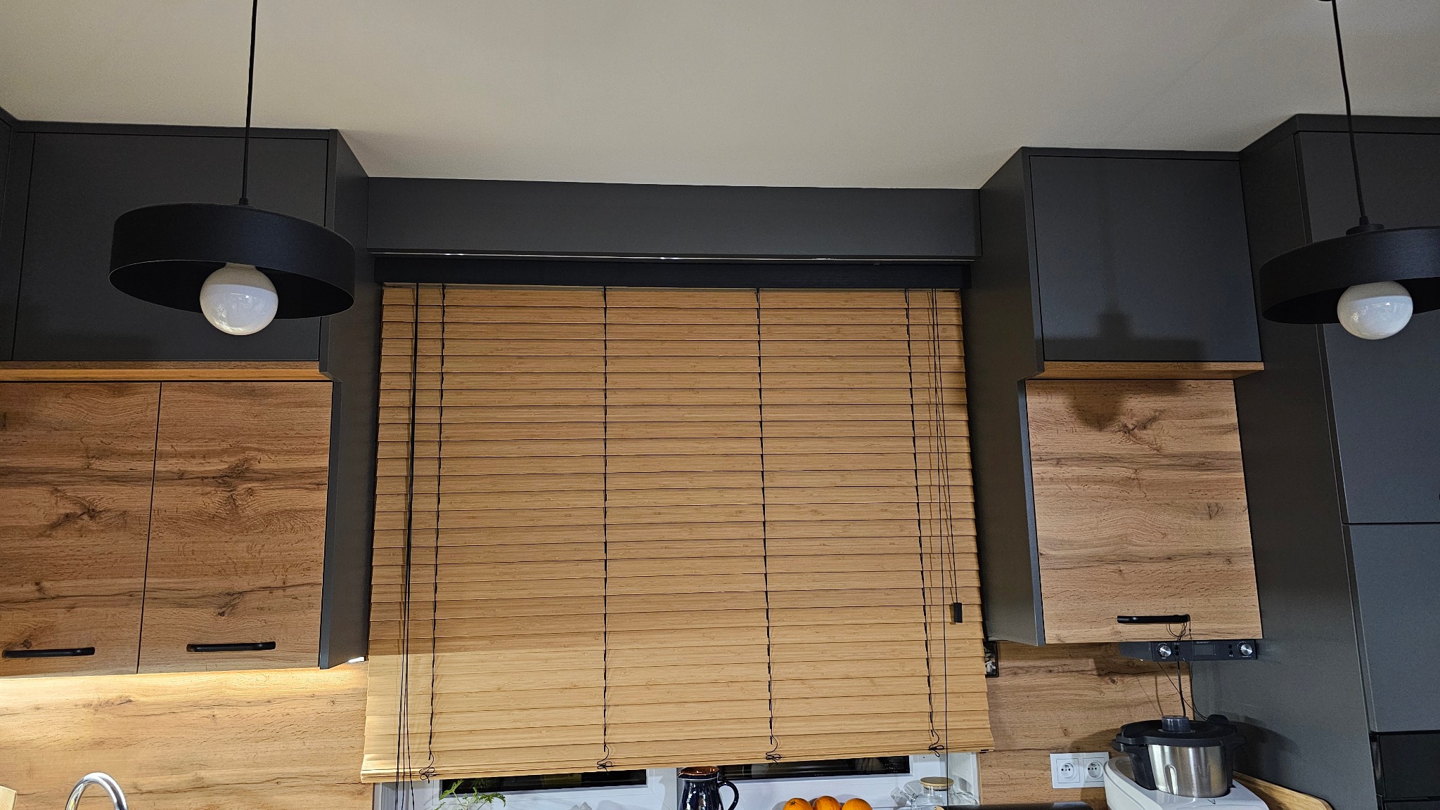 Jasne, poziome lamele w oknie kuchennym, drewniane szafki z widocznym usłojeniem, czarne lampy wiszące i ciemnoszare szafki górne tworzą spójną całość.