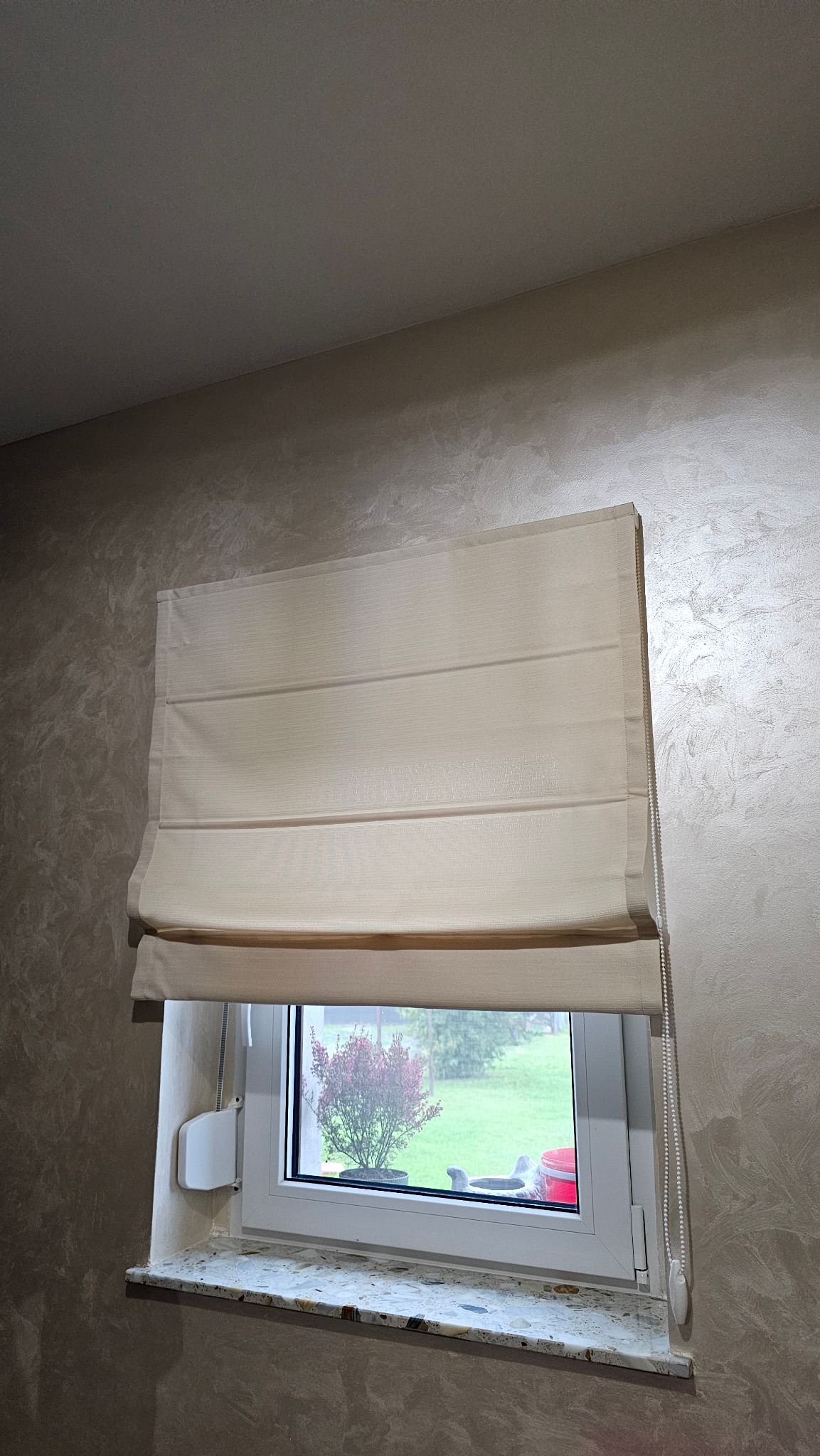 Beżowa, pozioma osłona okienna z łańcuszkiem, zamontowana w oknie z widokiem na zieleń. Dekoracyjna ściana w tle.