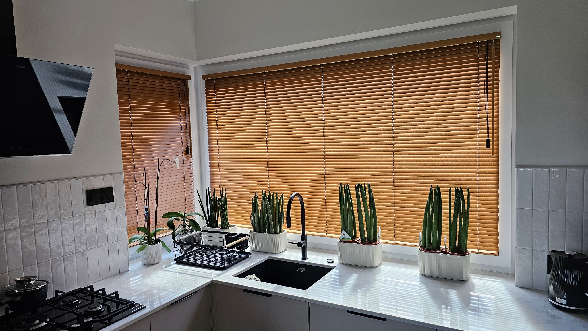 Kuchnia z oknem przesłoniętym poziomymi, drewnianymi lamelami. Na parapecie doniczki z zielonymi roślinami. Widok na zlew i płytę grzewczą.