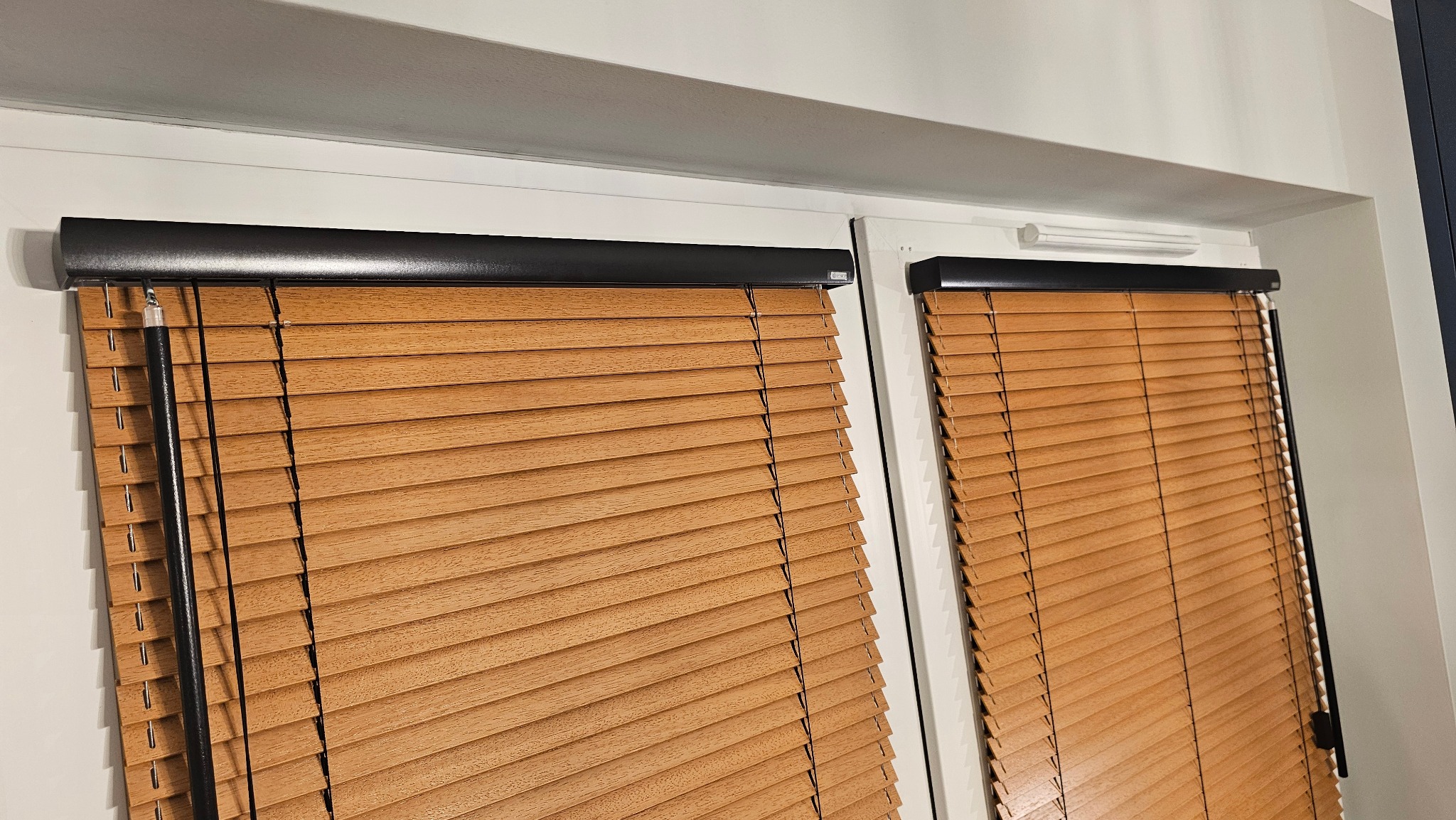 Dwa okna z zamontowanymi poziomymi, drewnianymi żaluzjami w odcieniach brązu, z czarnymi elementami mocującymi. Widok z bliska, wnętrze pomieszczenia.