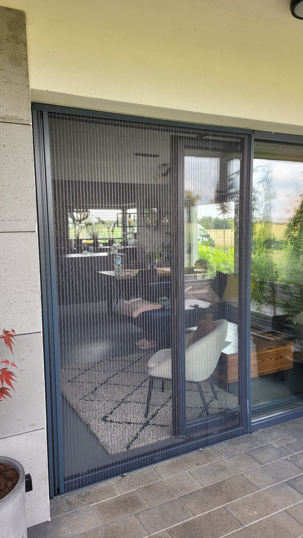 Zamknięta, przesuwna moskitiera plisowana w kolorze grafitowym, zamontowana na tarasowych drzwiach przesuwnych. Widok na wnętrze domu i ogród.
