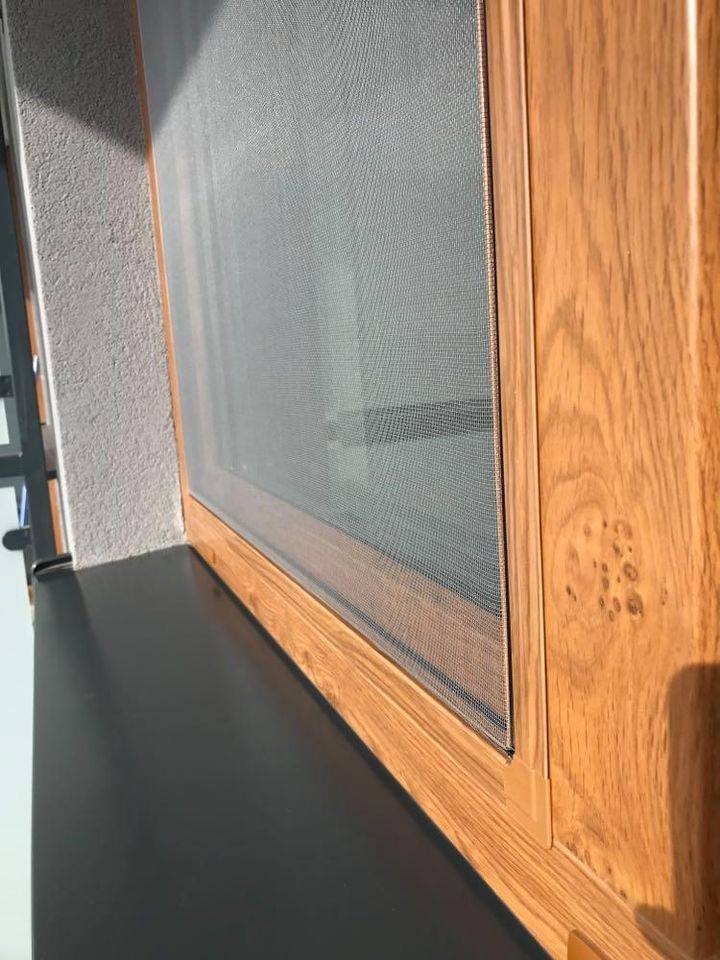 Zamontowana moskitiera w oknie z drewnianą ramą, widok z boku, szara ściana w tle. Siatka moskitiery jest napięta i równa.