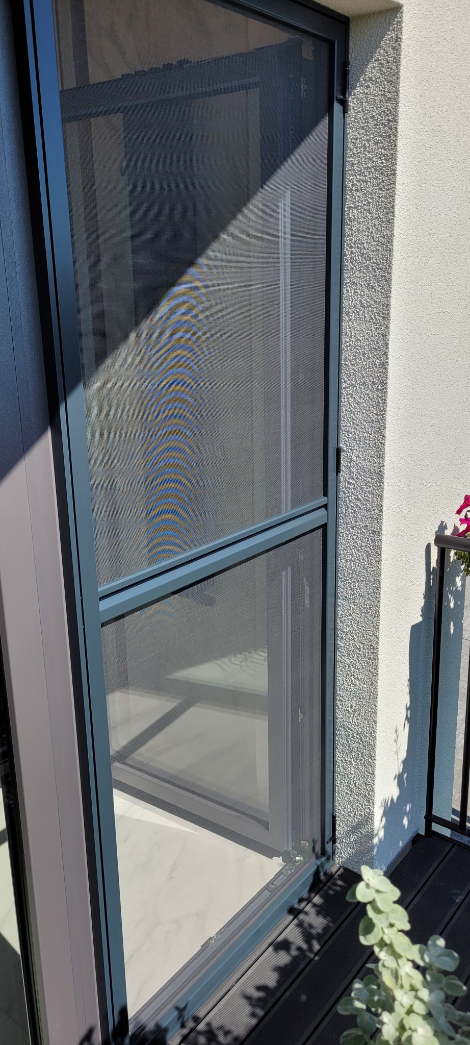 Szara moskitiera ramkowa w oknie, widok z balkonu. Na ścianie widoczne cienie, a w tle marmurowa podłoga. Dyskretna ochrona przed owadami.