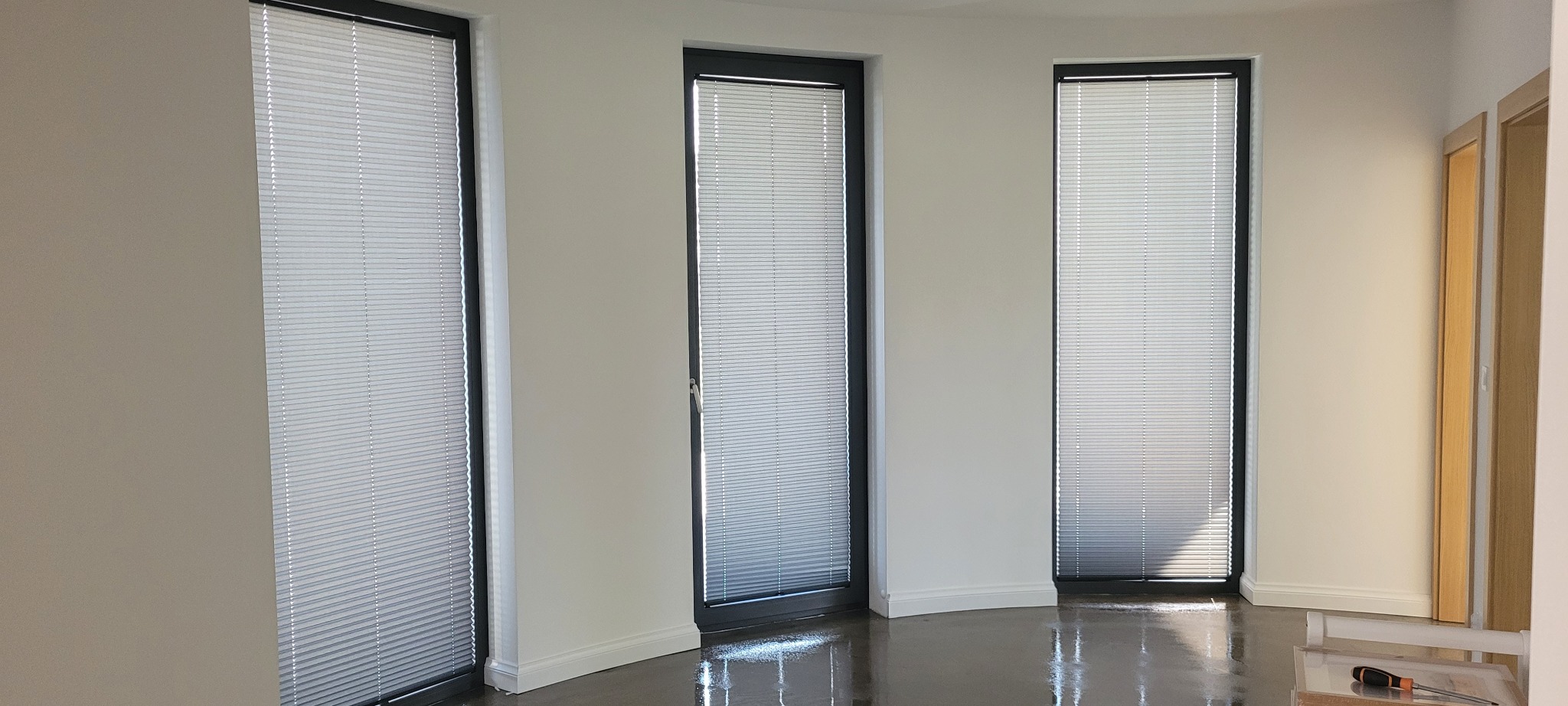 Trzy okna z zamontowanymi białymi plisami w nowoczesnym wnętrzu. Widoczny połysk podłogi i fragment narzędzi monterskich.