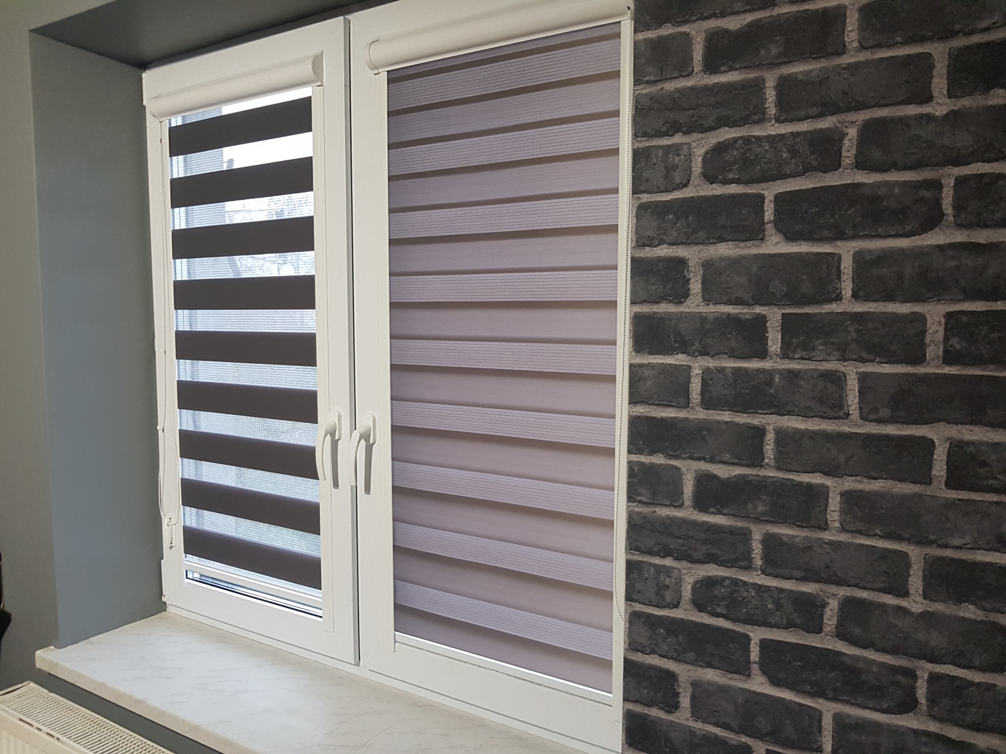 Dwa okna z zamontowanymi roletami dzień noc w kolorach szarości. Jedna roleta jest w pełni rozwinięta, druga częściowo zwinięta, ukazując efekt regulacji światła.