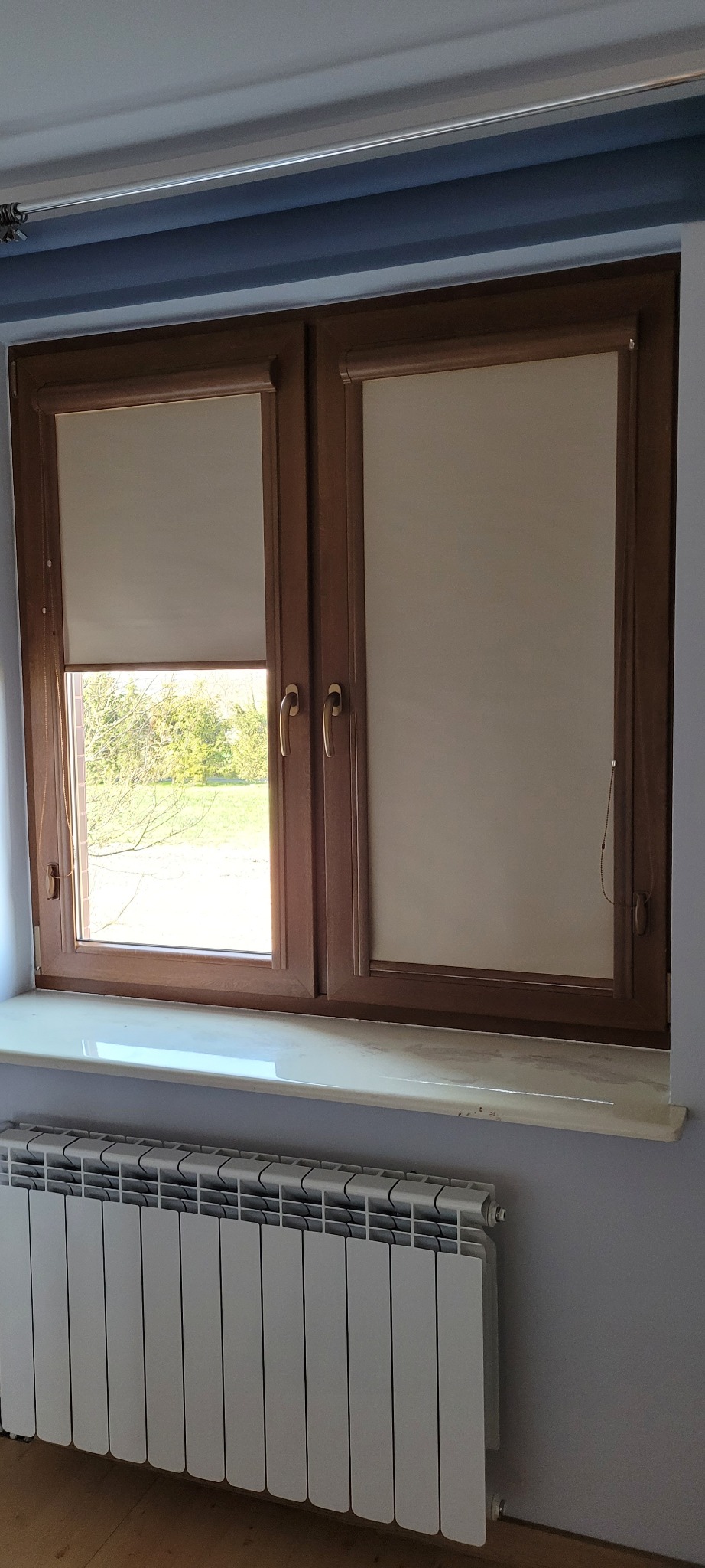 Dwuokienne okno z brązową ramą. Na oknach beżowe rolety materiałowe, jedna częściowo zwinięta, odsłaniająca widok na zieleń. Pod oknem biały grzejnik.