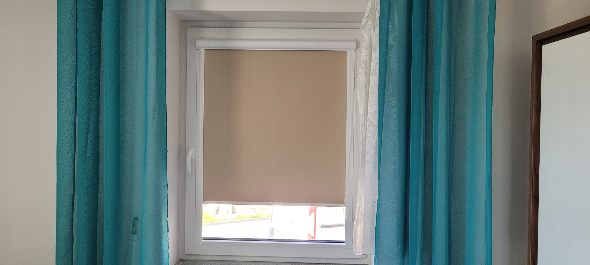 Beżowa roleta w oknie z turkusowymi zasłonami i firanką. Widok na zewnątrz. Minimalistyczny design, jasne wnętrze.