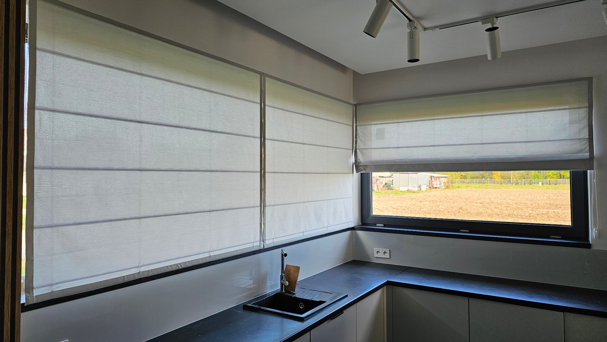 Jasne, poziome  fałdy rolet rzymskich w nowoczesnej kuchni z czarnym blatem i widokiem na pole. Minimalistyczny design, neutralna kolorystyka, równomierne oświetlenie.