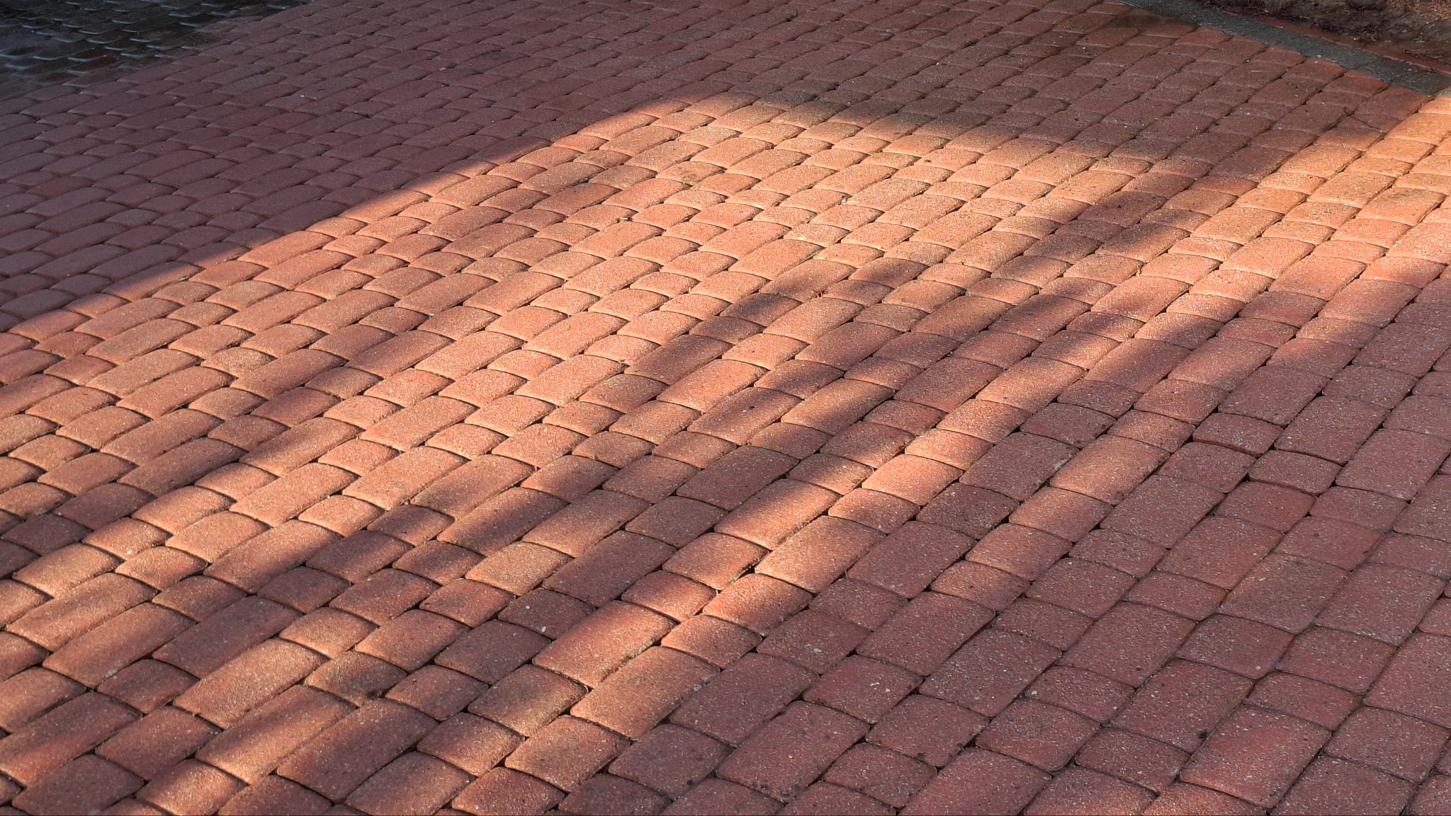 Czerwona kostka brukowa ułożona w jodełkę, oświetlona promieniami słońca tworzącymi cienie. Widok z góry, ukazujący regularny wzór i fakturę materiału.