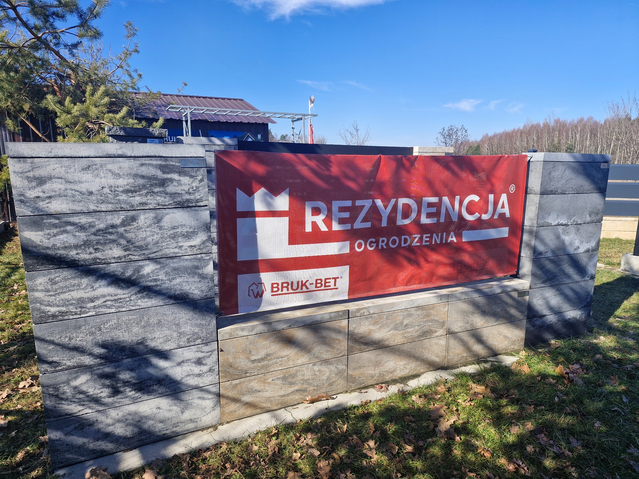 Nowoczesne ogrodzenie betonowe z szarego, melanżowego betonu, z czerwonym banerem reklamowym firmy Bruk-Bet 'Rezydecja Ogrodzenia' na tle zieleni i błękitnego nieba.