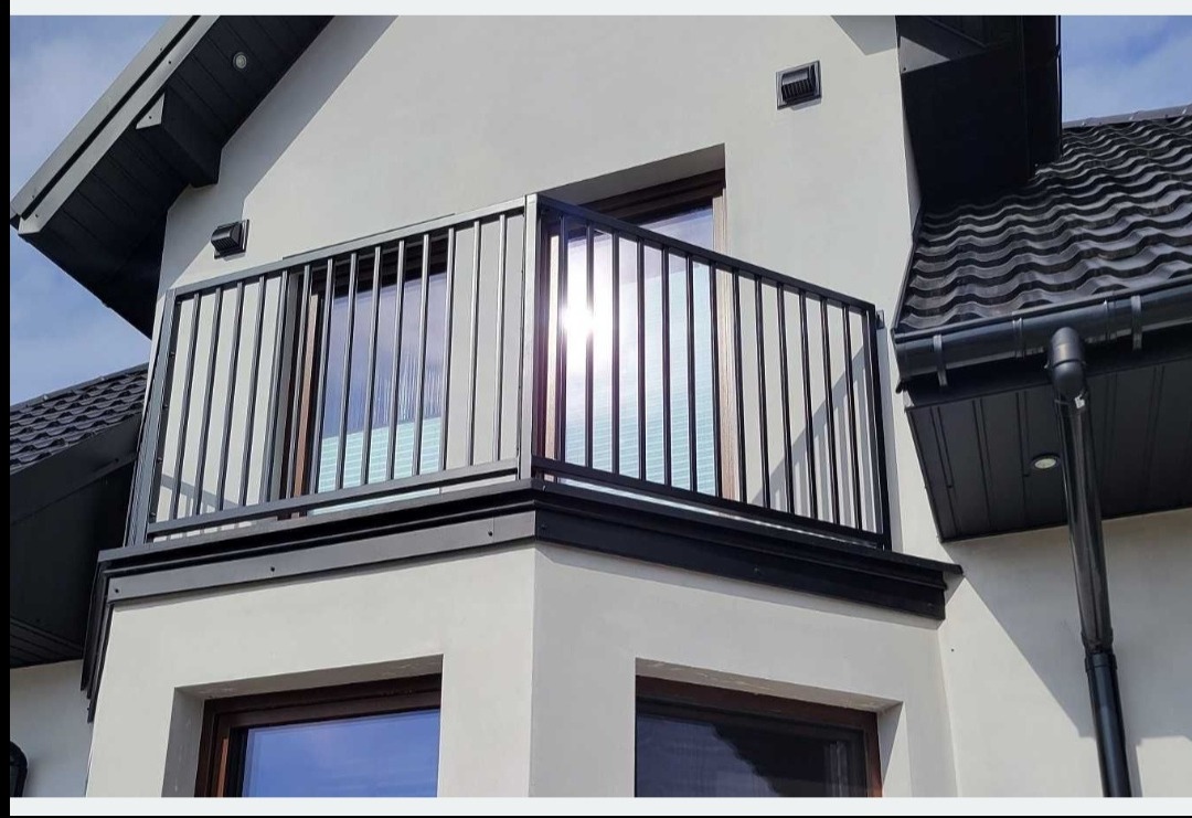 Nowoczesna, czarna balustrada balkonowa z pionowymi szczeblami, zamontowana na jasnym budynku z ciemnym dachem. Słońce odbija się w szybie okna za balustradą.