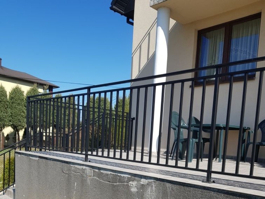 Czarna, metalowa balustrada balkonowa z pionowymi szczeblami, widok z zewnątrz, z zielonymi krzesłami i stołem na balkonie, w tle budynek z kolumną.