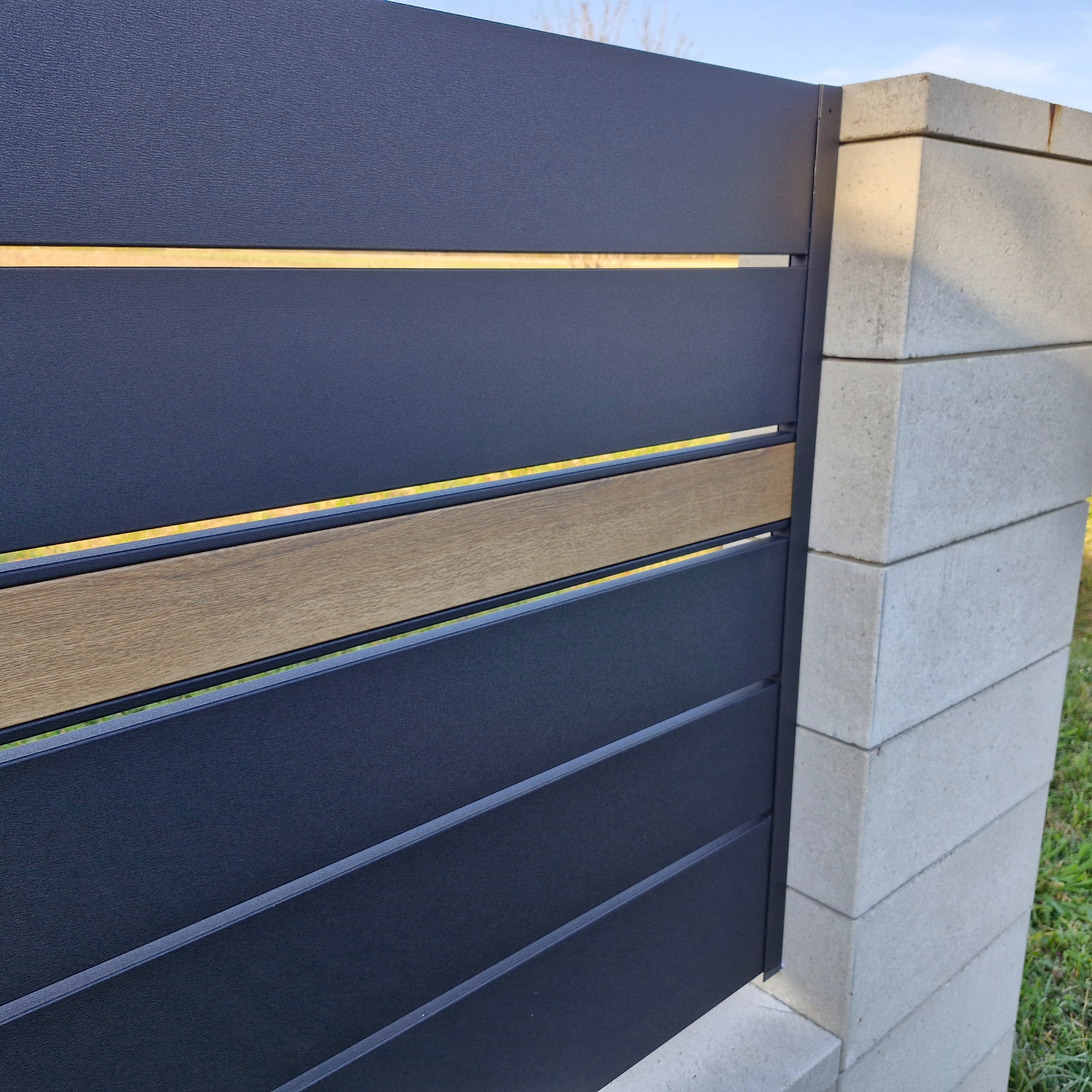 Nowoczesne ogrodzenie panelowe w kolorze grafitowym z poziomymi deskami imitującymi drewno, osadzone na słupkach z szarego betonu architektonicznego.