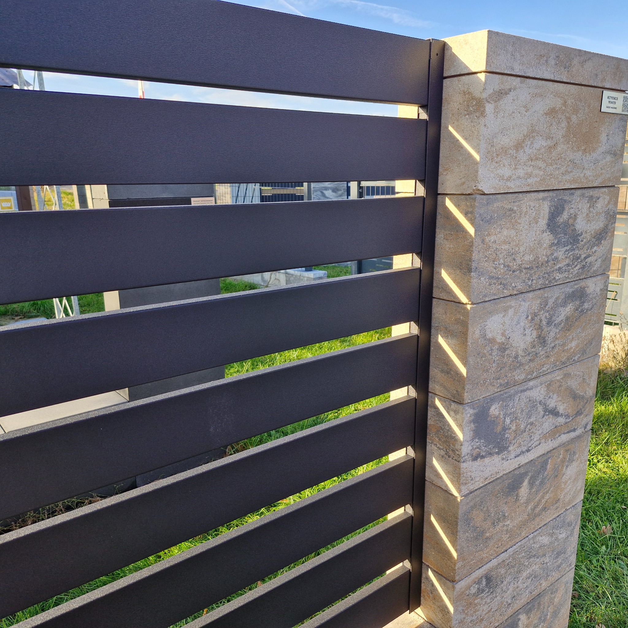 Nowoczesne ogrodzenie panelowe z poziomych, grafitowych listew, wsparte na słupkach z beżowo-szarego kamienia. Widok z bliska na fragment ogrodzenia.