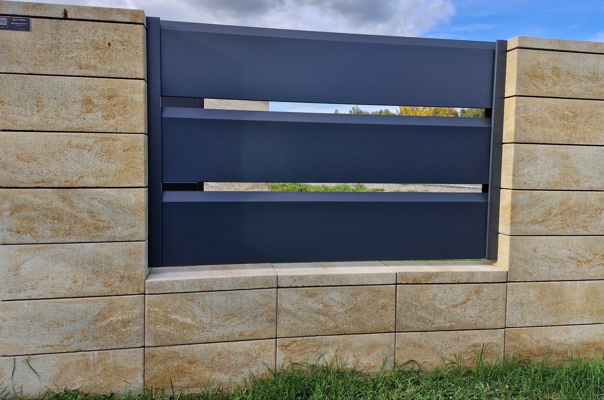 Nowoczesne ogrodzenie: grafitowe, poziome panele metalowe między słupkami z beżowych, warstwowych bloczków. Widok z poziomu trawy, błękitne niebo w tle.