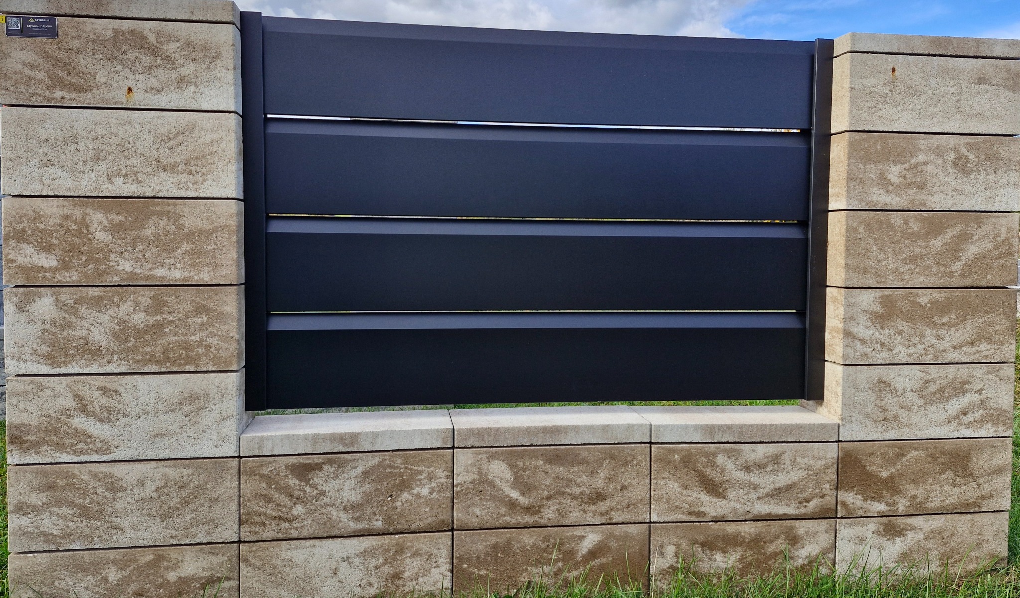 Nowoczesne ogrodzenie: czarne, poziome panele metalowe z subtelnymi, srebrnymi akcentami, osadzone na słupkach z beżowych, teksturowanych bloczków betonowych.