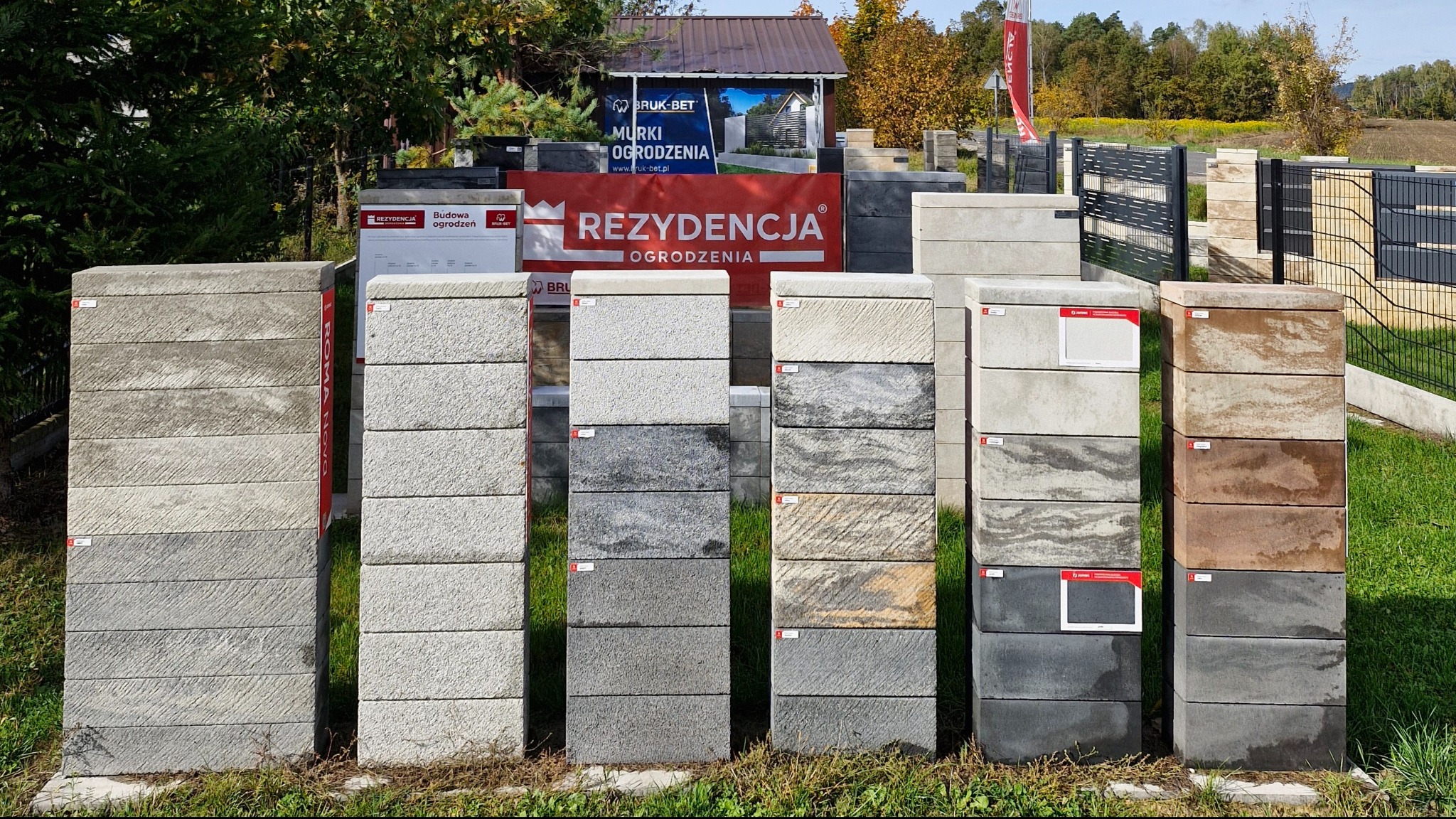 Ekspozycja różnorodnych bloczków betonowych imitujących kamień, ustawionych w kolumny na trawie, prezentacja kolorów i faktur. W tle ogrodzenie panelowe.