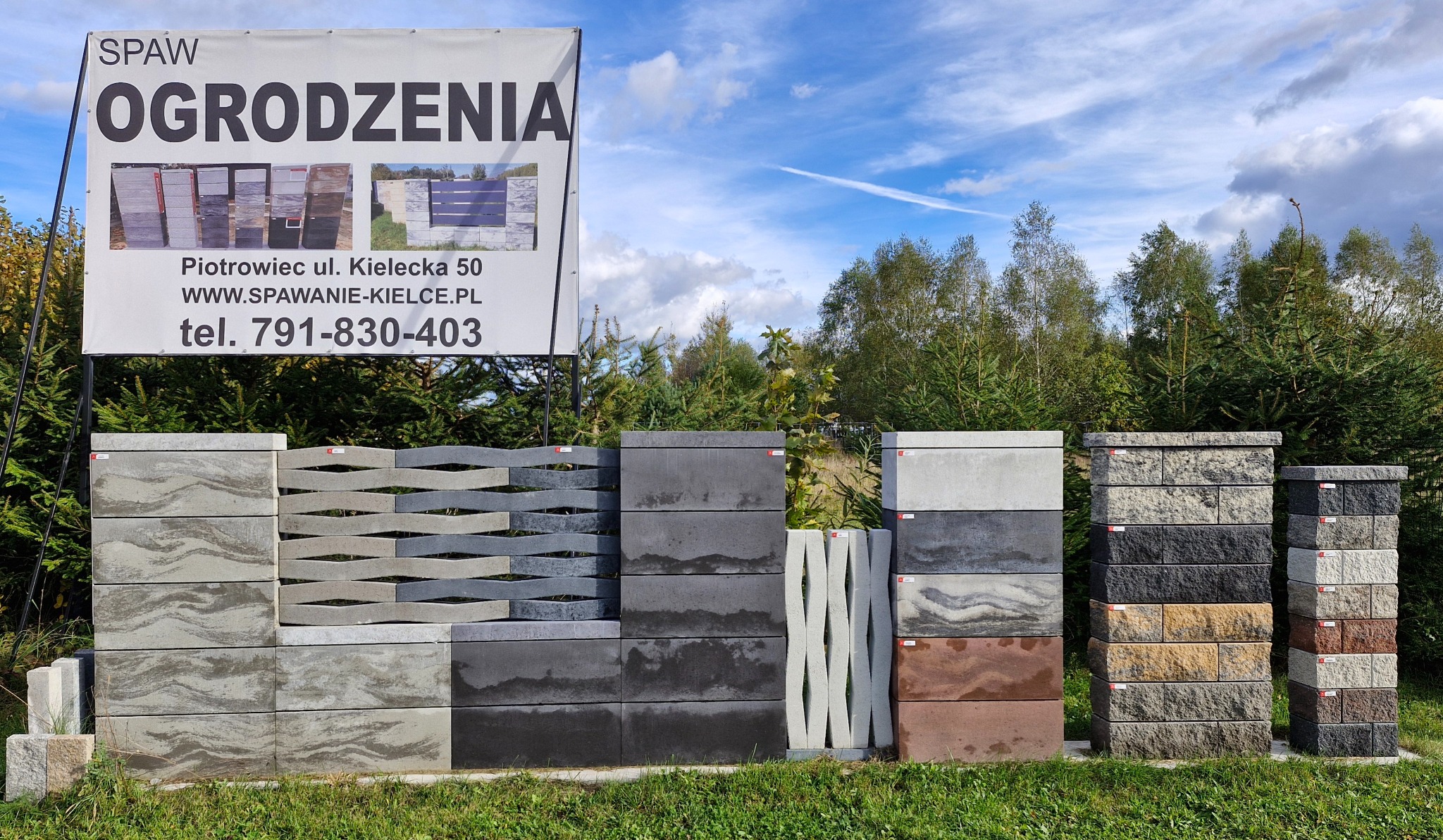 Ekspozycja różnorodnych bloczków ogrodzeniowych z betonu, prezentowanych na tle zieleni i błękitnego nieba. Widoczny baner reklamowy firmy z danymi kontaktowymi.