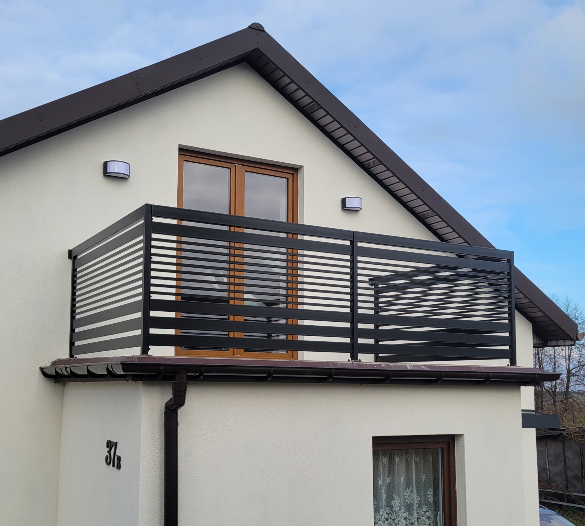 Nowoczesna, czarna balustrada balkonowa z poziomych profili na tle białej elewacji domu z brązowym dachem i oknami. Widok z dołu, błękitne niebo w tle.