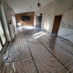 Piotr Sikora - Białe rury ogrzewania podłogowego ułożone na folii z czerwoną kratką w remontowanym pomieszczeniu. Widoczne okna, grzejnik i fragmenty ścian w stanie surowym.