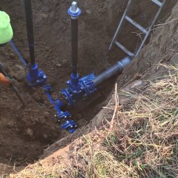 Piotr Sikora - Niebieska instalacja wodociągowa w wykopie ziemnym, widoczna drabina i fragment łopaty z ziemią. Prace ziemne i instalacyjne w toku.