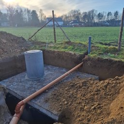 Piotr Sikora - Betonowy zbiornik z rurą kanalizacyjną w wykopie ziemnym na tle pola i zabudowań. Widoczne elementy instalacji przyłącza wod-kan w trakcie montażu.