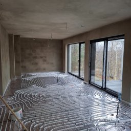 Piotr Sikora - Białe rury ogrzewania podłogowego ułożone na folii w surowym wnętrzu z dużymi oknami. Wiadro i szczotka w rogu kadru.