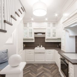 Biała kuchnia pod schodami z drewnianym blatem i podłogą w jodełkę. Uwagę przykuwa ceramiczny zlew farmerski i dekoracyjne szafki z przeszkleniami.