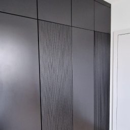 Czarna szafa wnękowa z ryflowanymi frontami, widok z dołu, podkreślający wysokość zabudowy i minimalistyczny design. Jasna podłoga i białe ściany kontrastują z ciemnym meblem.