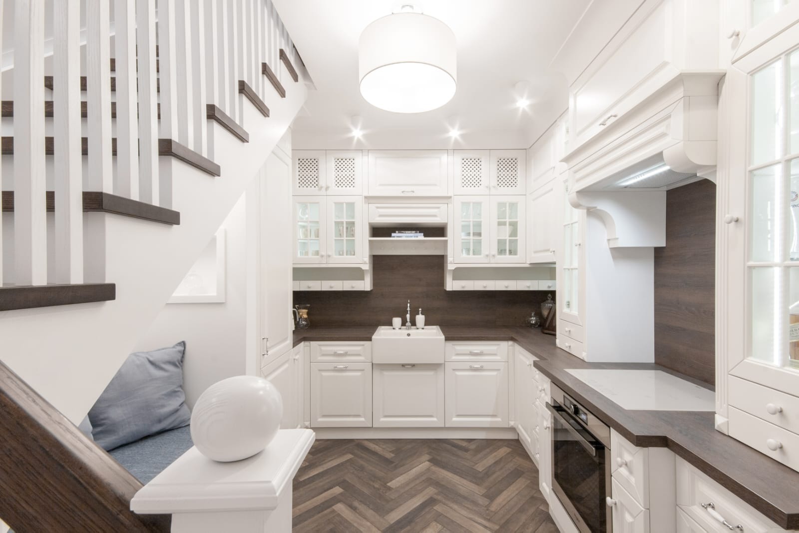 Biała kuchnia pod schodami z drewnianym blatem i podłogą w jodełkę. Uwagę przykuwa ceramiczny zlew farmerski i dekoracyjne szafki z przeszkleniami.