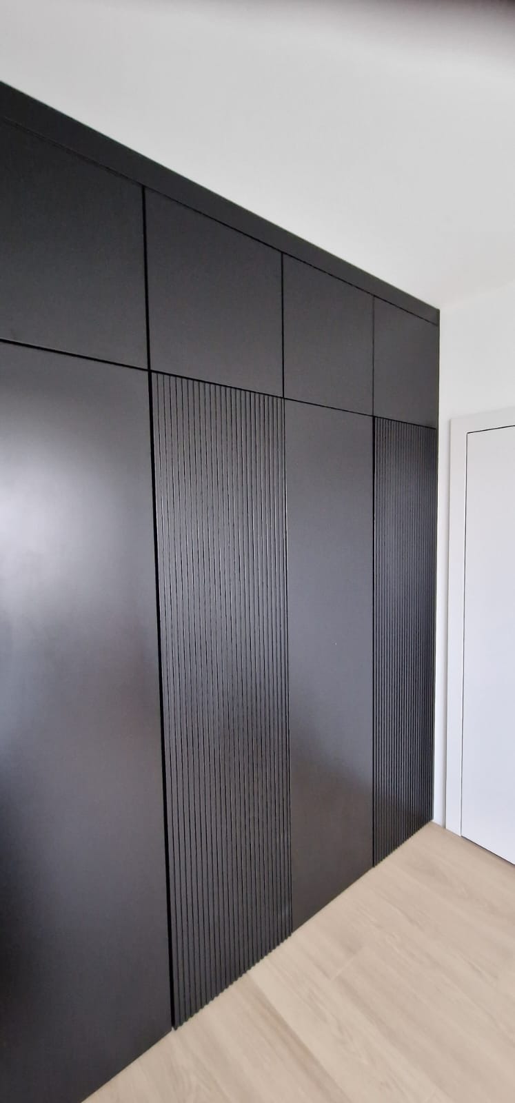 Czarna szafa wnękowa z ryflowanymi frontami, widok z dołu, podkreślający wysokość zabudowy i minimalistyczny design. Jasna podłoga i białe ściany kontrastują z ciemnym meblem.