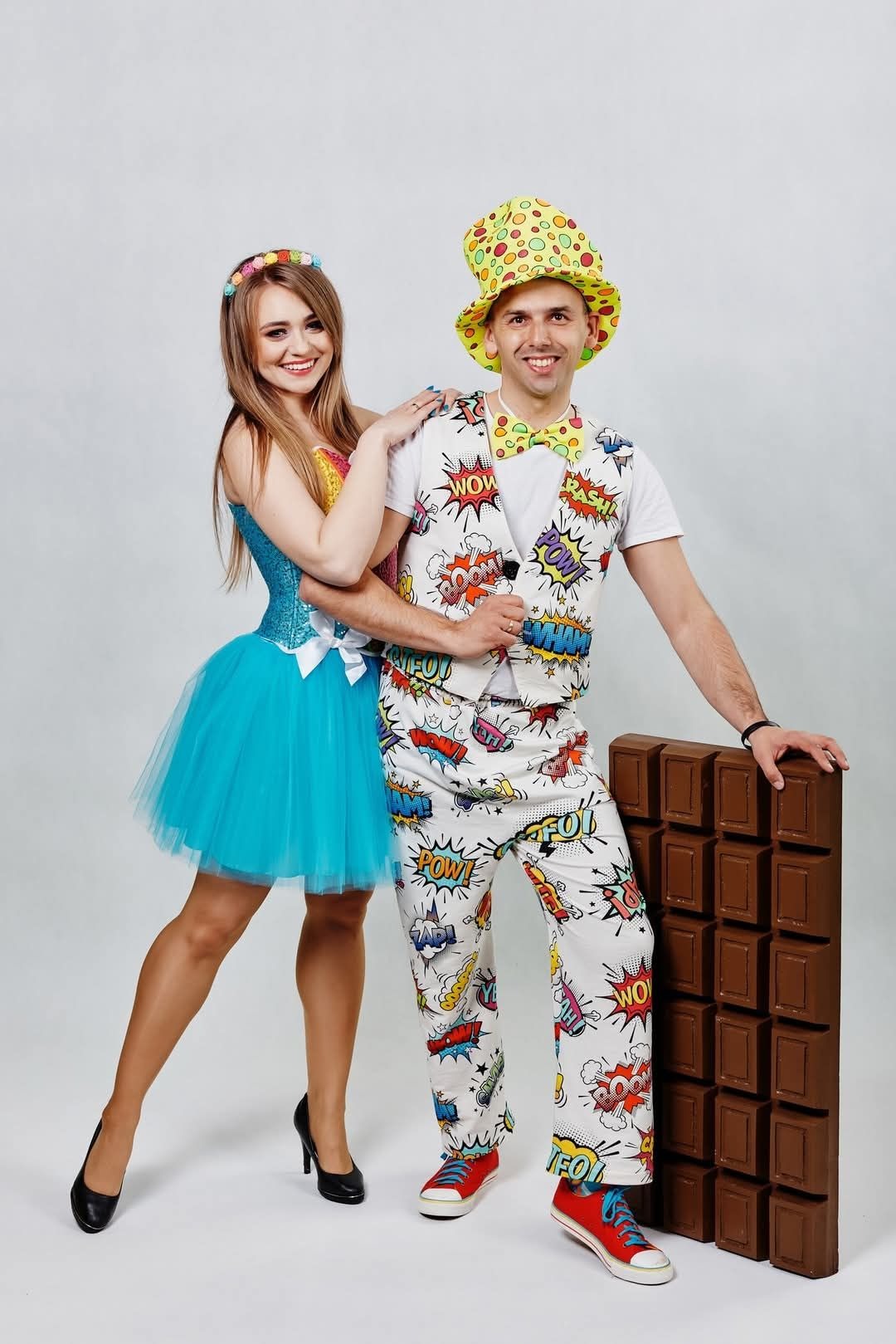 Uśmiechnięci animatorzy w kostiumach komiksowych, pozujący obok rekwizytu imitującego tabliczkę czekolady na jasnym tle. Dziewczyna w niebieskiej sukience, mężczyzna w kapeluszu.