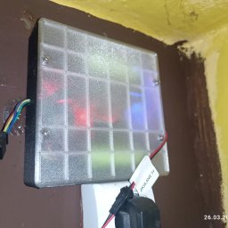 CADexo ADAM SKALIK - Prostopadłościenna obudowa z przezroczystego tworzywa, przymocowana do ściany, z widocznymi przewodami i elementami elektronicznymi wewnątrz. Widoczne ślady montażu.