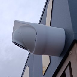 CADexo ADAM SKALIK - Zewnętrzna kamera monitoringu, zamontowana na elewacji budynku o nowoczesnym designie. Widoczne elementy drewniane i metalowe. Ujęcie z dołu, pochmurne niebo w tle.