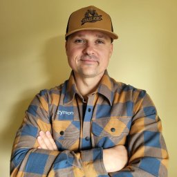 S&S Logistics Szymon Siemieniako - Uśmiechnięty mężczyzna w czapce Strauss Works i koszuli w kratę z logo 'zymon' na tle jednolitej ściany. Skrzyżowane ręce, portret.