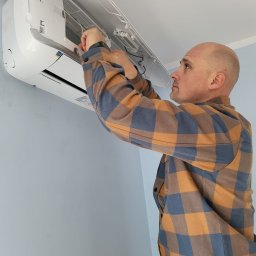 S&S Logistics Szymon Siemieniako - Mężczyzna w kraciastej koszuli instaluje klimatyzator na jasnej ścianie. Widoczny otwarty panel urządzenia i dłoń montera przy pracy.