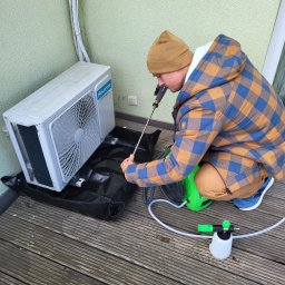 S&S Logistics Szymon Siemieniako - Czyszczenie jednostki zewnętrznej klimatyzacji Hisense na balkonie. Mężczyzna w czapce i kurtce w kratę używa lancy i myjki ciśnieniowej. Czarna mata pod urządzeniem.