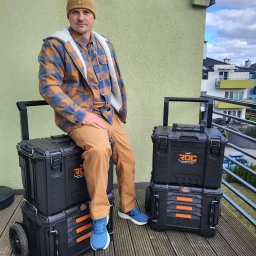 Pingwin klimatyzacja - Mężczyzna w czapce i flanelowej koszuli siedzi na czarnych skrzynkach narzędziowych ROC na kółkach, na tle balkonu i budynku mieszkalnego w słoneczny dzień.
