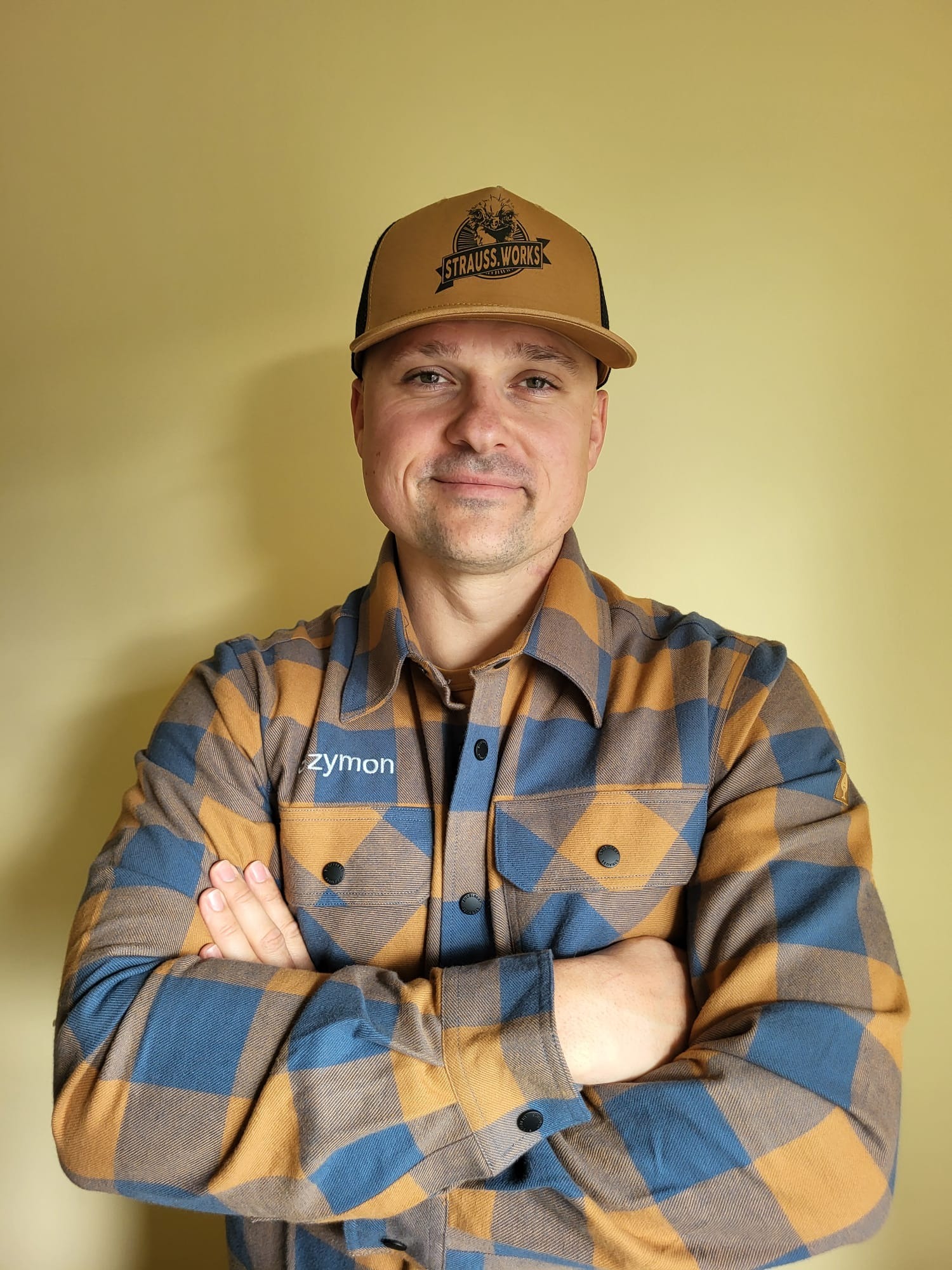 Uśmiechnięty mężczyzna w czapce Strauss Works i koszuli w kratę z logo 'zymon' na tle jednolitej ściany. Skrzyżowane ręce, portret.