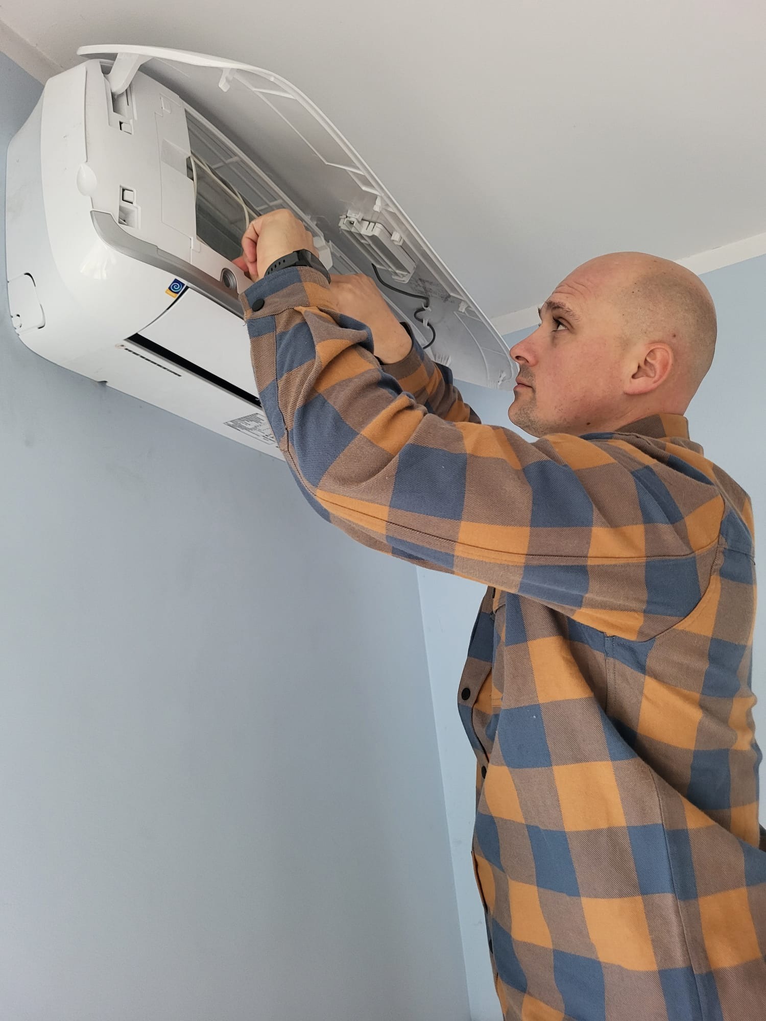 Mężczyzna w kraciastej koszuli instaluje klimatyzator na jasnej ścianie. Widoczny otwarty panel urządzenia i dłoń montera przy pracy.