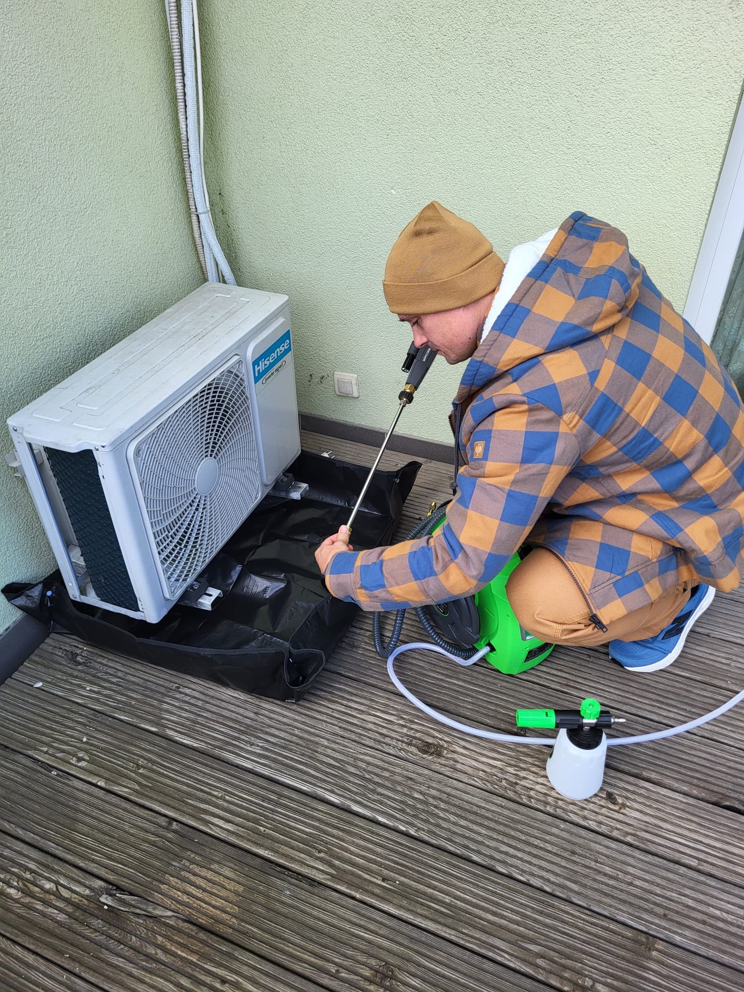Czyszczenie jednostki zewnętrznej klimatyzacji Hisense na balkonie. Mężczyzna w czapce i kurtce w kratę używa lancy i myjki ciśnieniowej. Czarna mata pod urządzeniem.