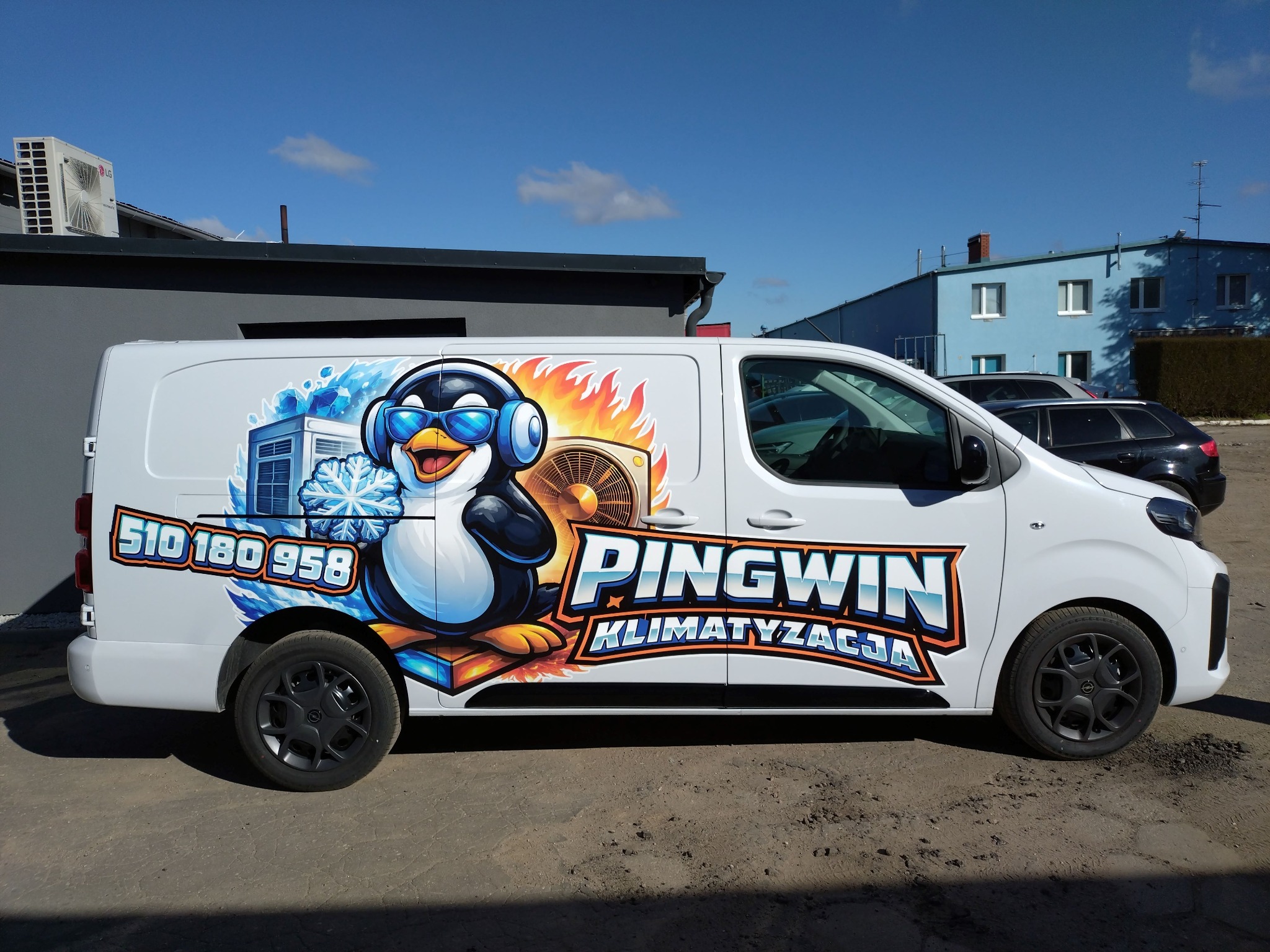 Biały samochód dostawczy z grafiką pingwina w okularach i słuchawkach, logo firmy 'Pingwin Klimatyzacja' i numerem telefonu na boku, na tle budynku z klimatyzatorem.