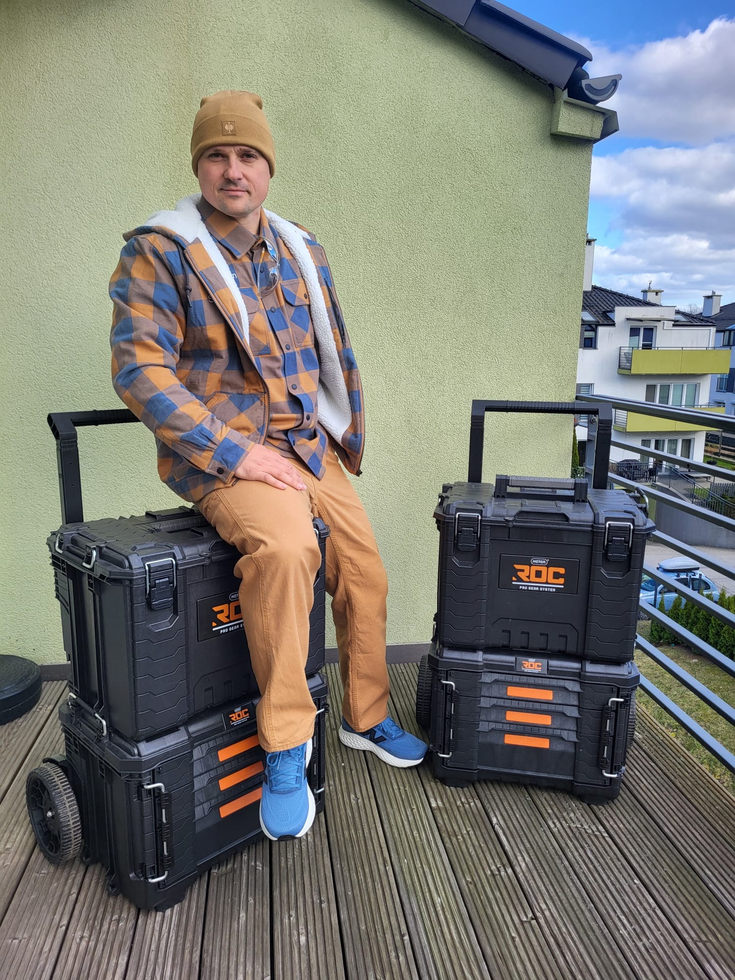 Mężczyzna w czapce i flanelowej koszuli siedzi na czarnych skrzynkach narzędziowych ROC na kółkach, na tle balkonu i budynku mieszkalnego w słoneczny dzień.